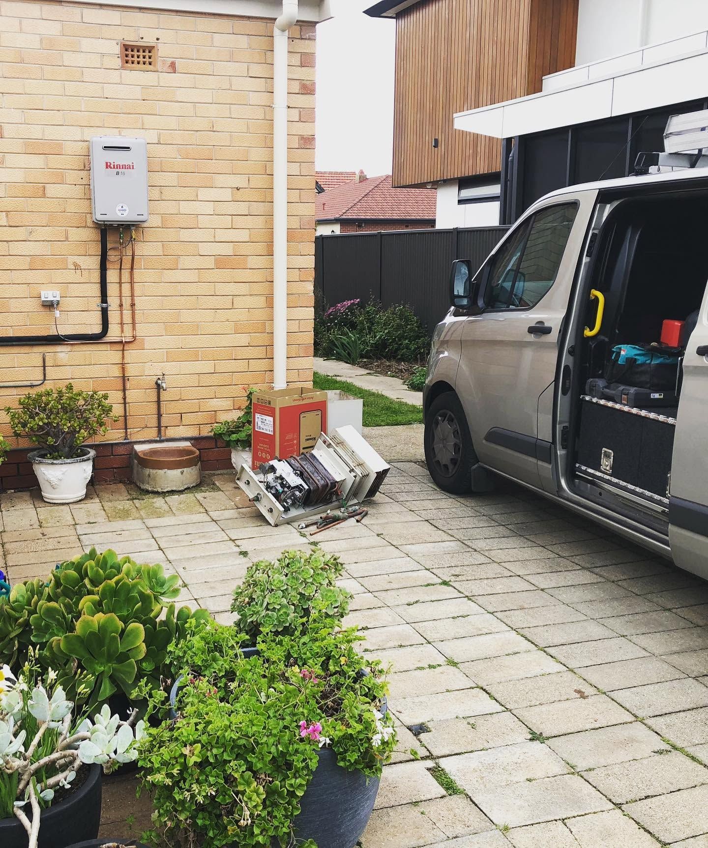 White van parked by brick house with potted plants and garden items on a paved driveway.