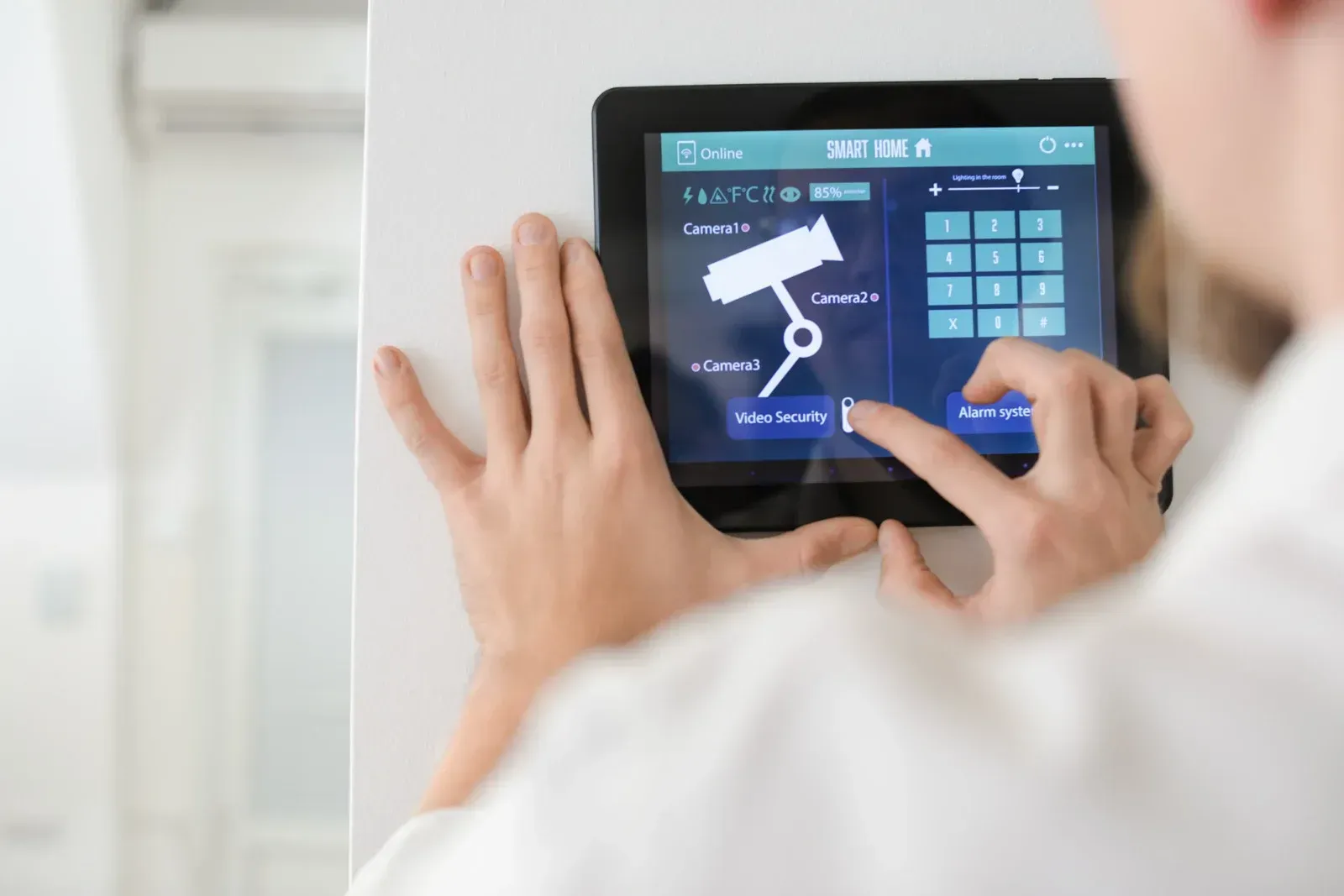 Person using a tablet on a wall, possibly for security system control. Blue UI, keypad, and camera icon visible.