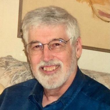 Smiling man with gray hair, glasses, and a beard, wearing a blue shirt on a couch