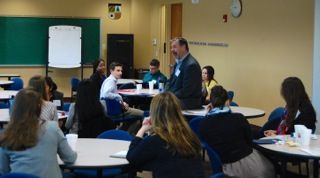 People seated in a classroom while a man speaks near a whiteboard at the front.