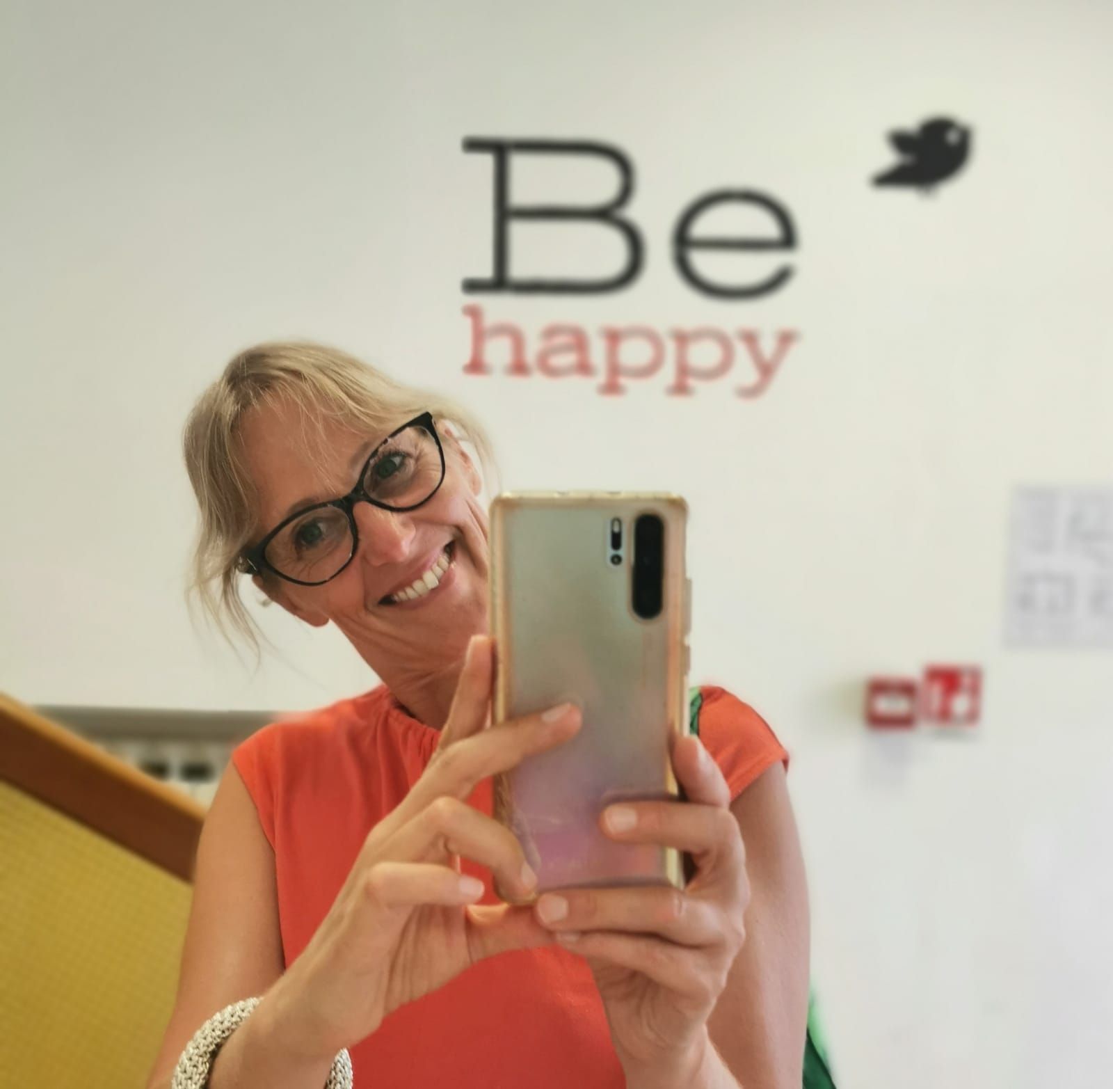 Donna che si scatta un selfie, sorride, tiene in mano il telefono e ha la scritta murale 