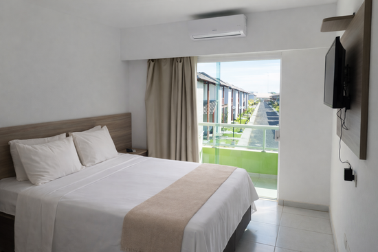 Quarto de hotel com decoração em tons de branco e bege, duas camas, uma pequena TV e uma varanda com vegetação.