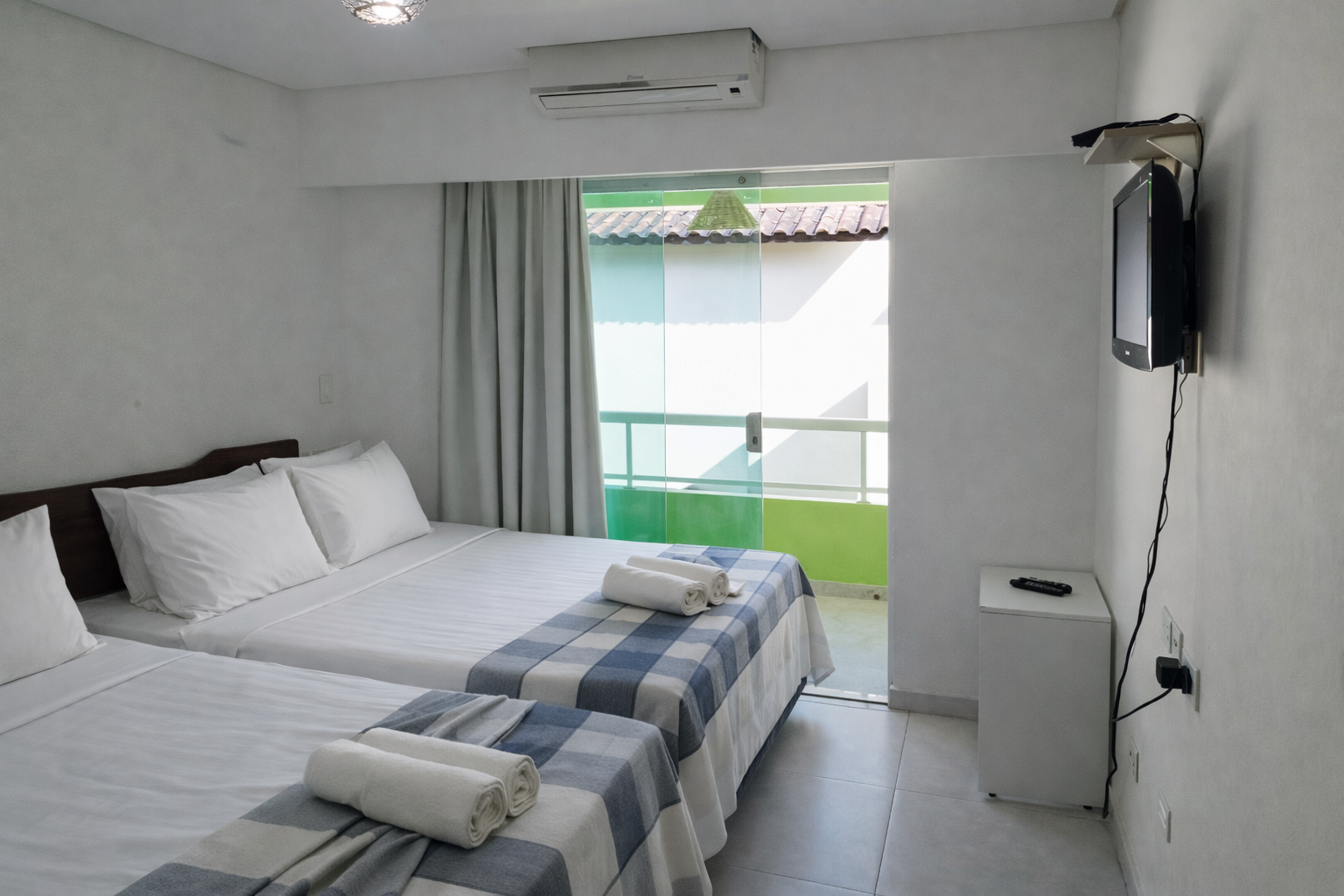 Quarto com duas camas, paredes brancas, varanda, TV e ar-condicionado. Toalhas e colchas azuis e brancas.