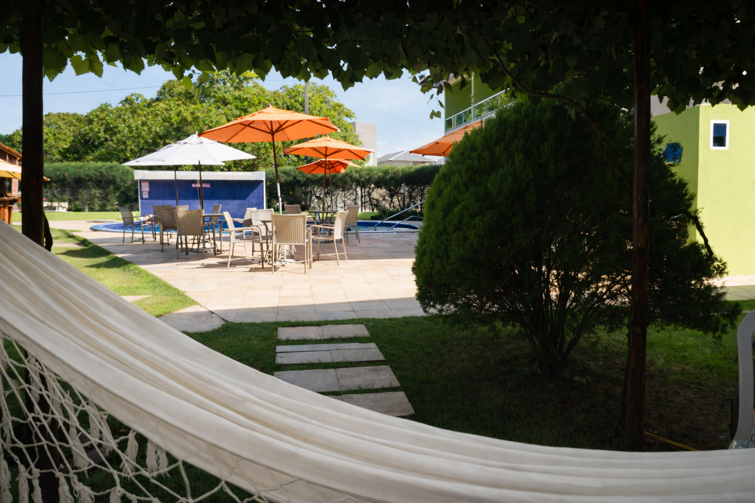 Gramado com rede, mesas, guarda-sóis e área de piscina sob as árvores.