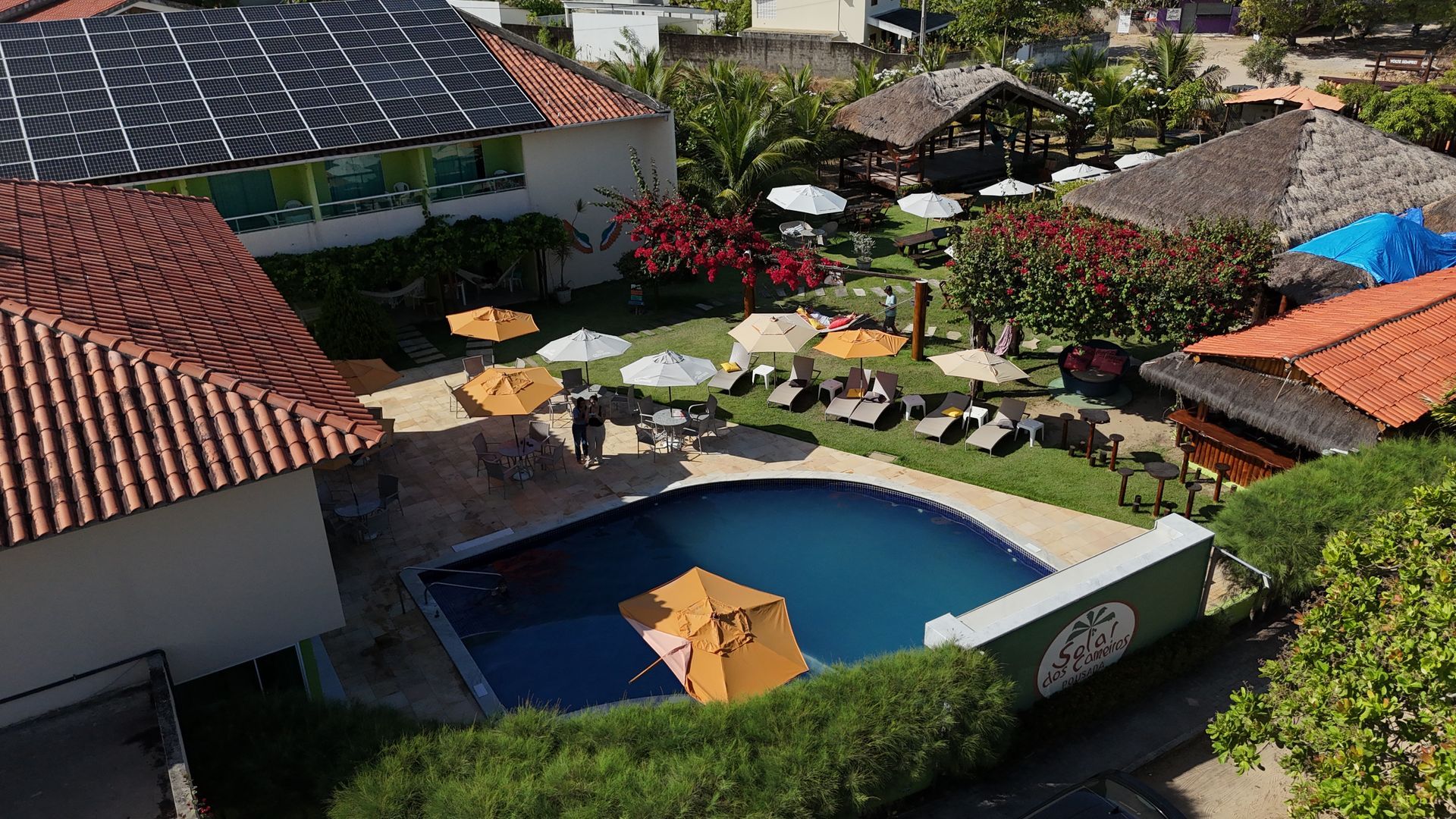 Vista aérea de um resort com piscina, espreguiçadeiras, guarda-sóis e edifícios com telhados vermelhos e pretos.