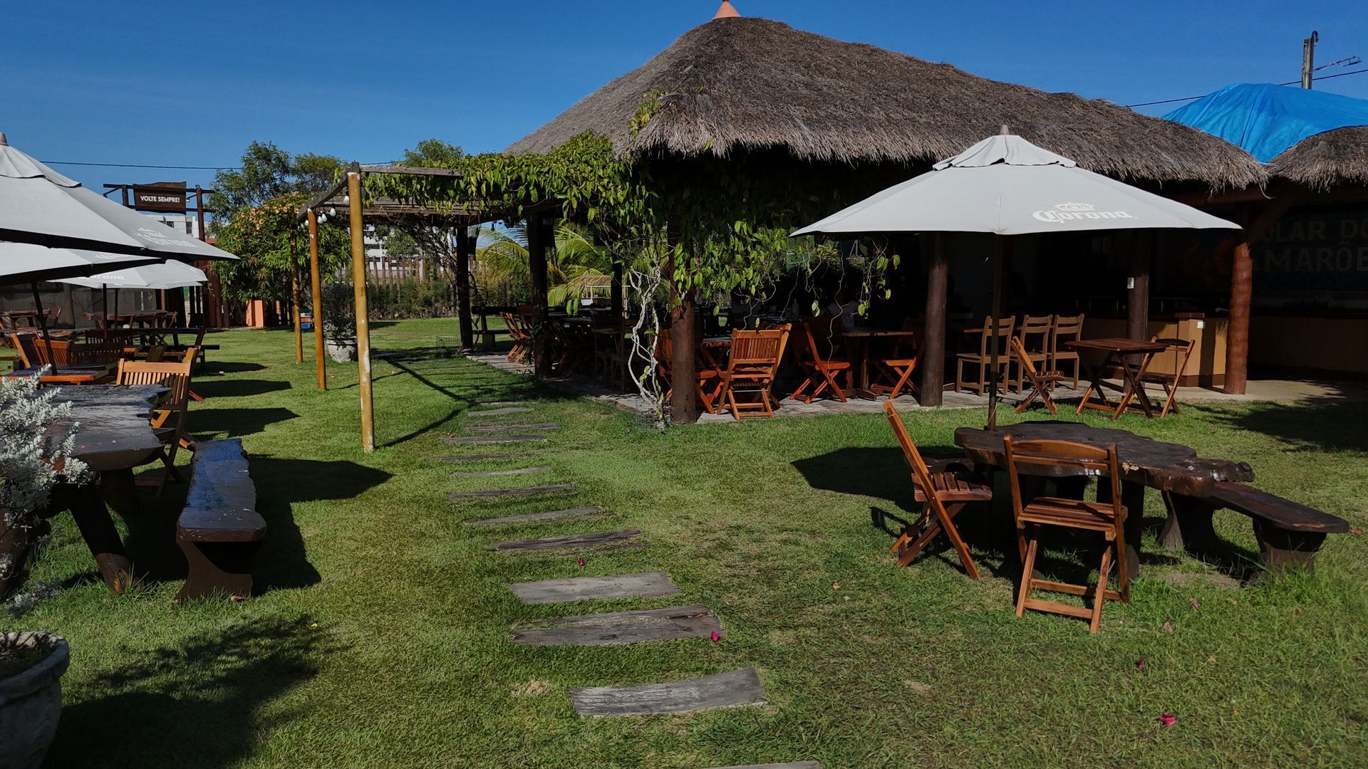 Restaurante ao ar livre com estrutura de telhado de palha, mesas de madeira e grama verde.