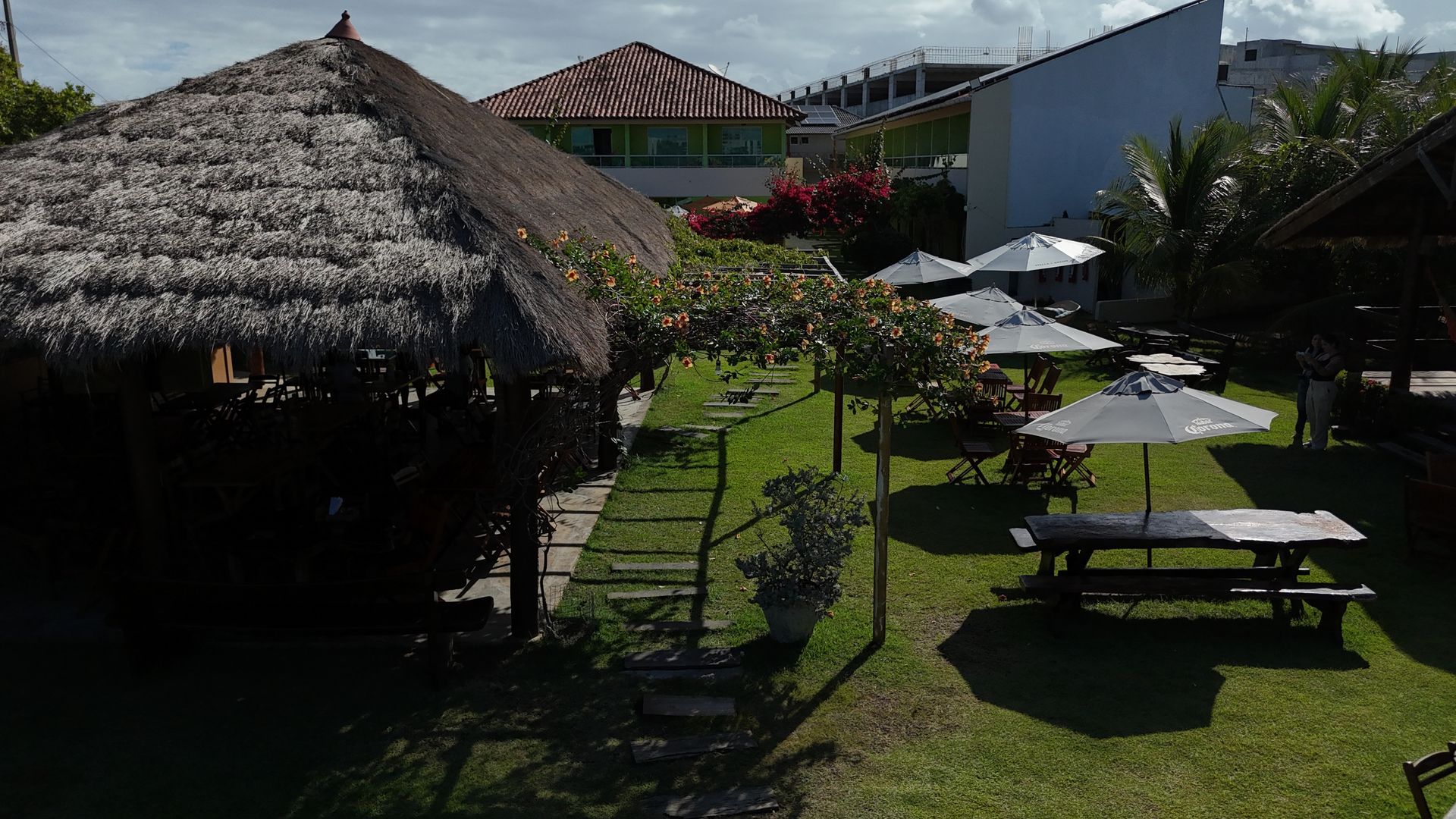 Quintal verdejante com estrutura de telhado de palha e mesas de piquenique com guarda-sóis.