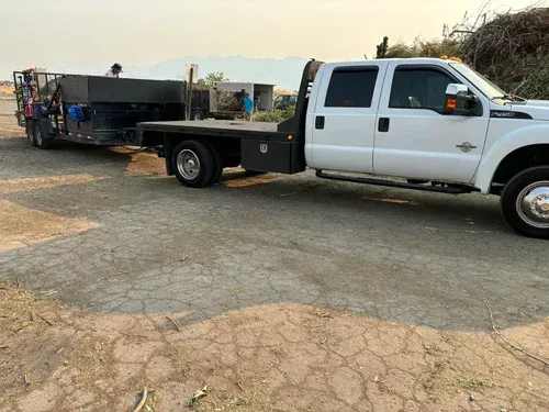 White pickup truck towing a trailer with machinery on a dirt road, mountains in background.