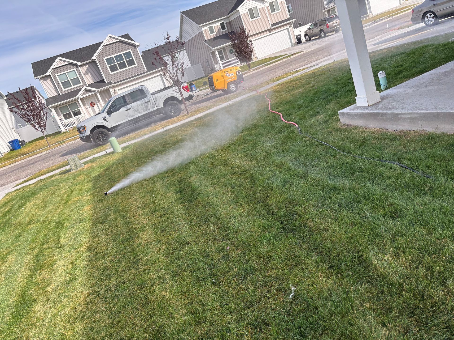 Sprinkler watering a green lawn in front of a house, a truck and other vehicles on the street.