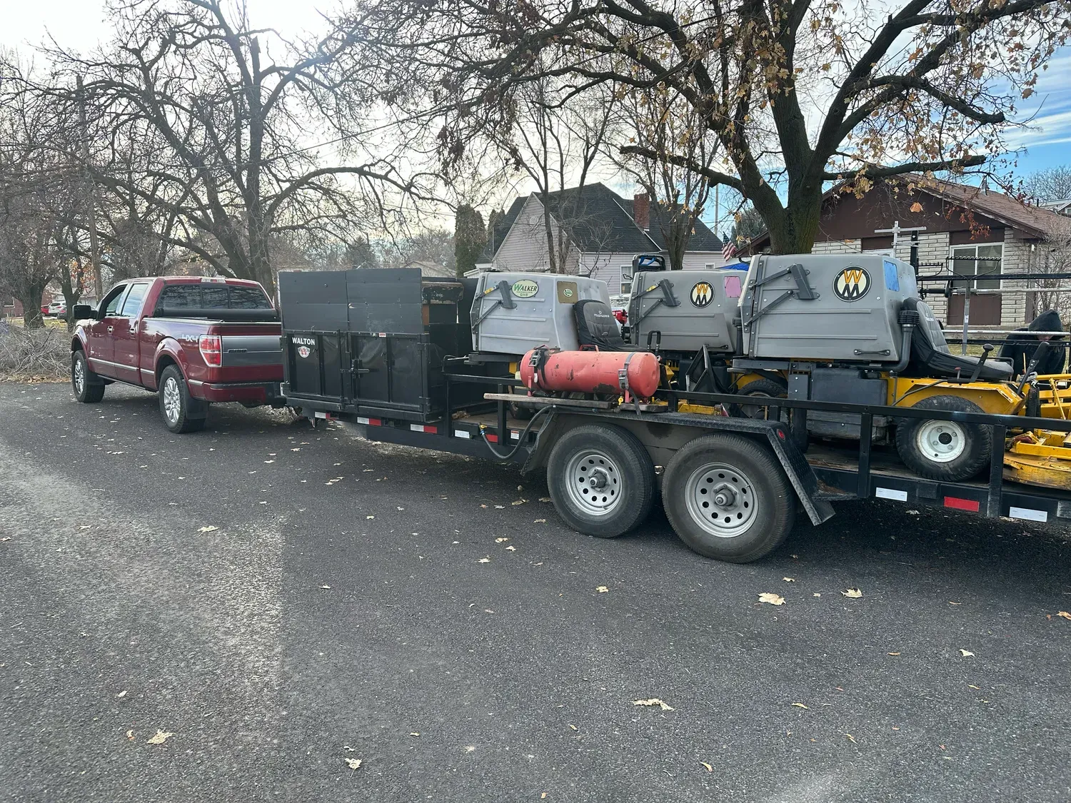 Red truck towing a trailer with landscaping equipment and a gray dumpster in a residential area.