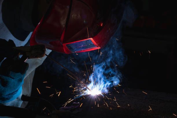 Welder using welding torch on metal with sparks and protective helmet in industrial workshop.