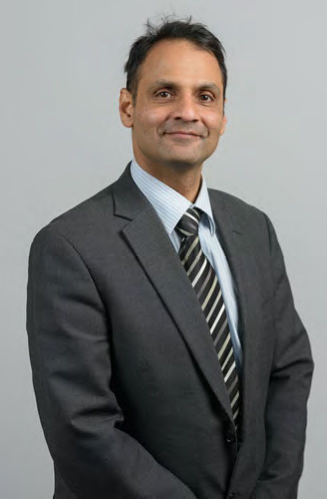 Man in a dark suit and striped tie smiles slightly against a gray background.