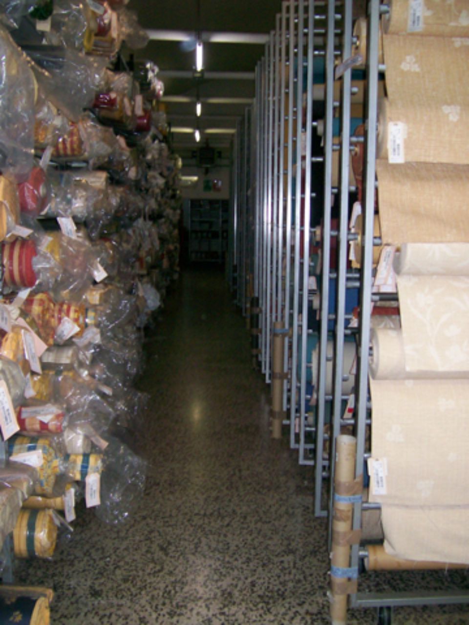 Scaffali pieni di rotoli di tessuto in un magazzino, lungo corridoio con scaffalature metalliche.