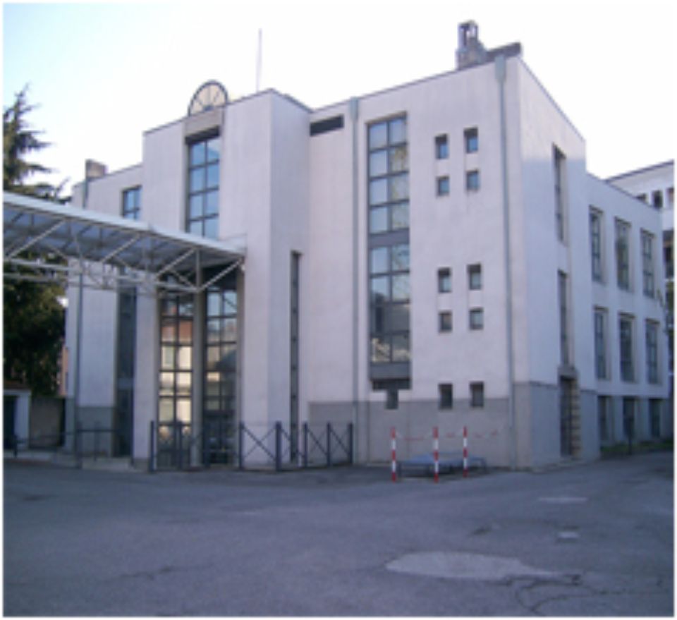 Edificio bianco con ampie finestre, ingresso coperto e area recintata.