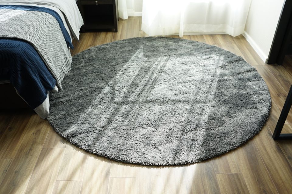Tappeto rotondo grigio sul pavimento in legno, vicino a un letto con una coperta blu. La luce del sole filtra a fiotti.