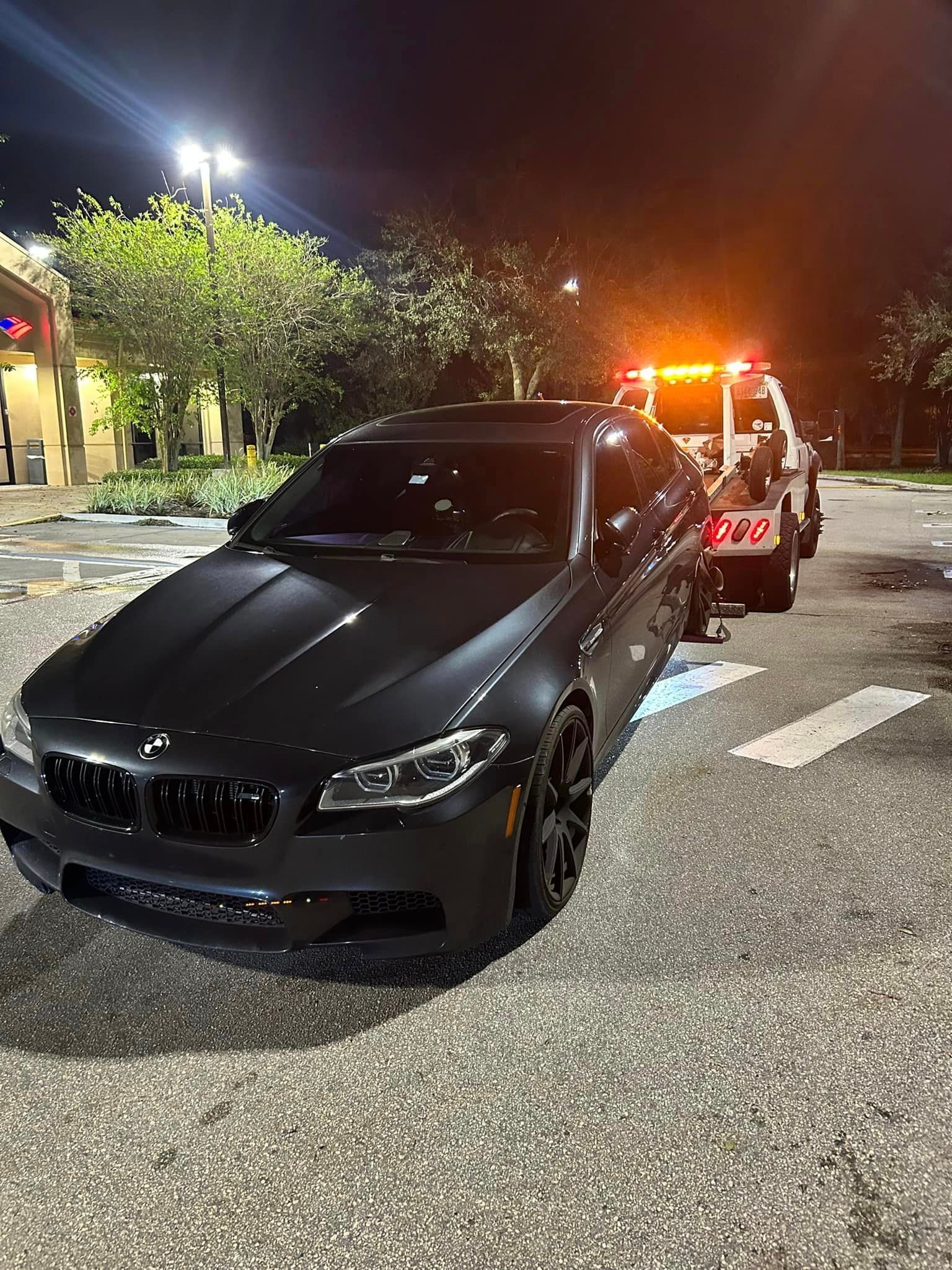 A black car is being towed by a tow truck at night.