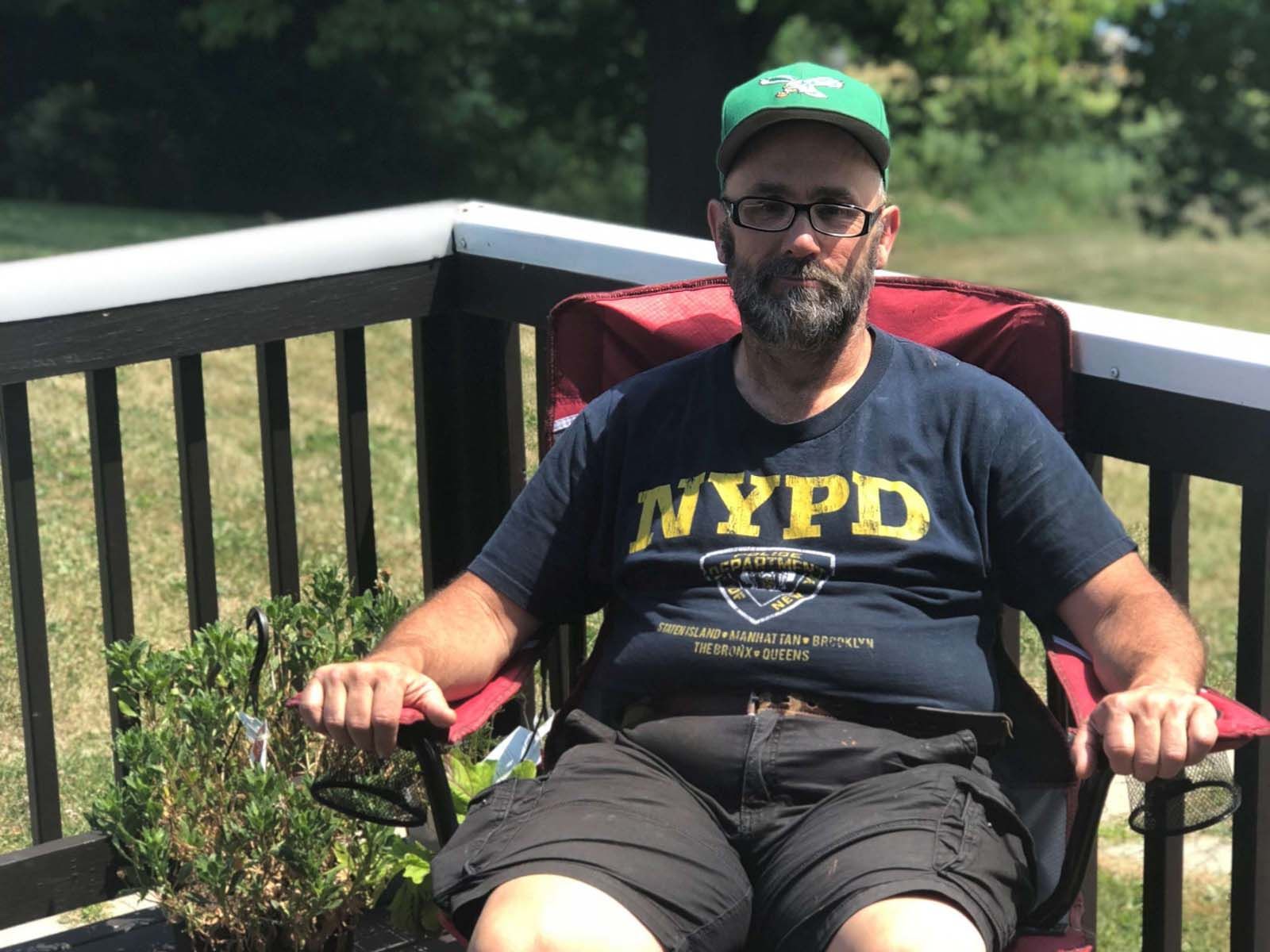 Person seated outdoors in a folding chair on a wooden deck, wearing a dark graphic T-shirt, shorts, glasses, and a green cap, with grass and trees visible in the background.