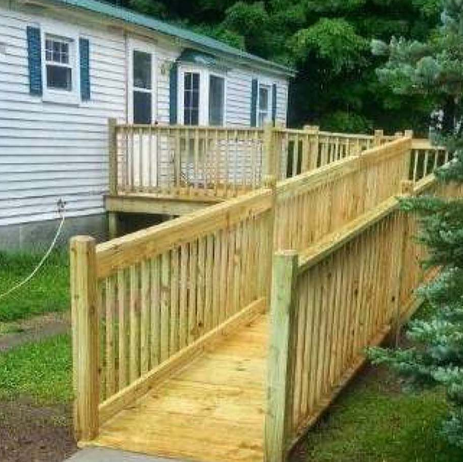 Exterior view of a single-story house with a wooden accessibility ramp and railings leading to a raised entrance, surrounded by grass and trees.