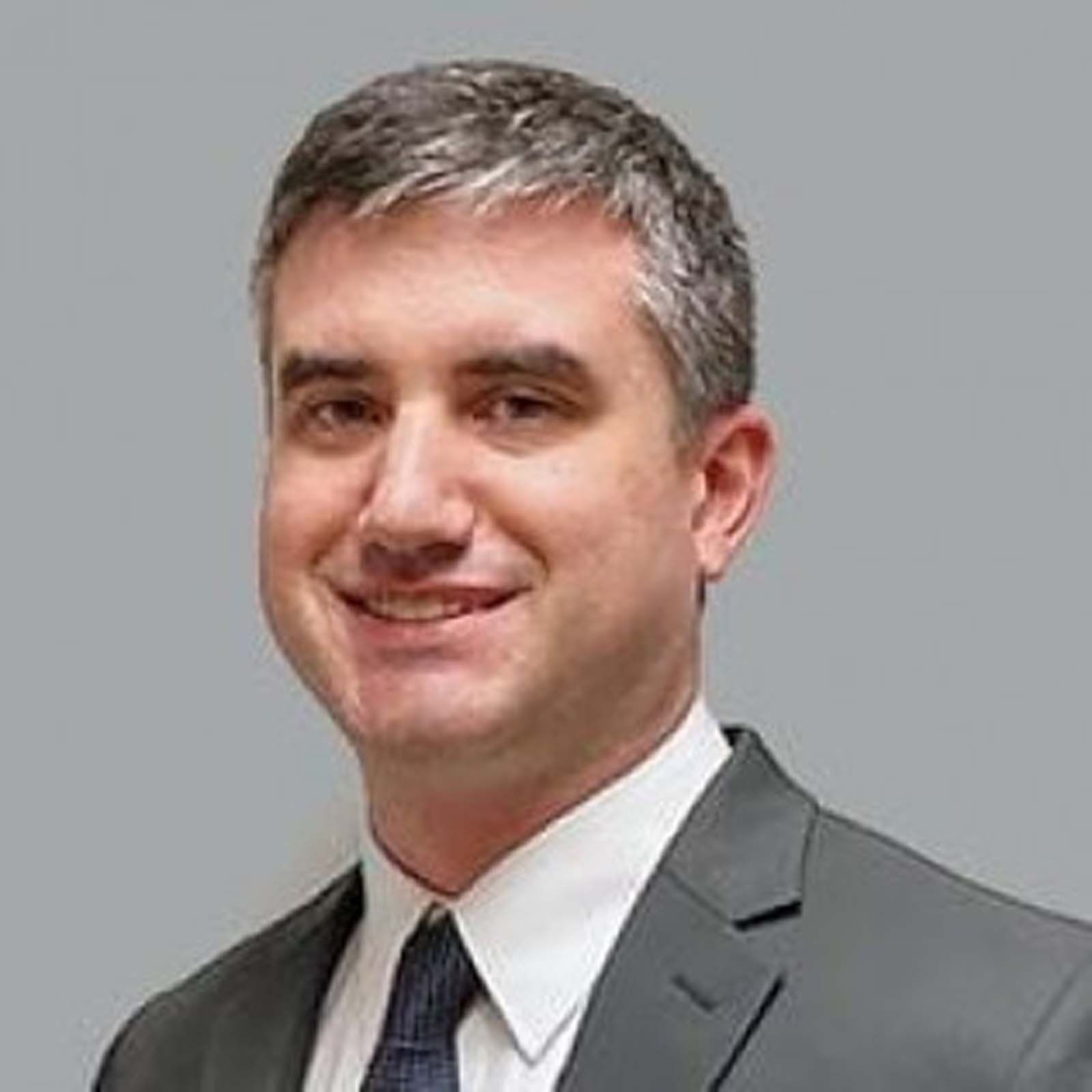 Portrait of a professional staff member wearing a dark suit jacket, white dress shirt, and dark tie, standing against a plain light-colored background.