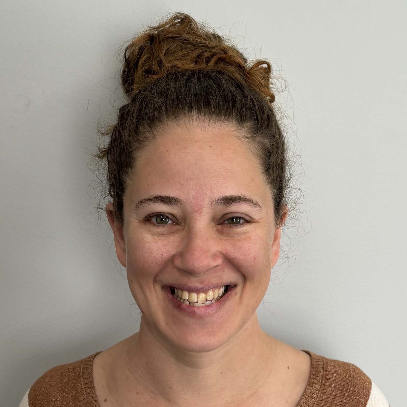Portrait of a professional staff member with hair pulled back, wearing a brown-and-white top, standing against a plain light colored background.