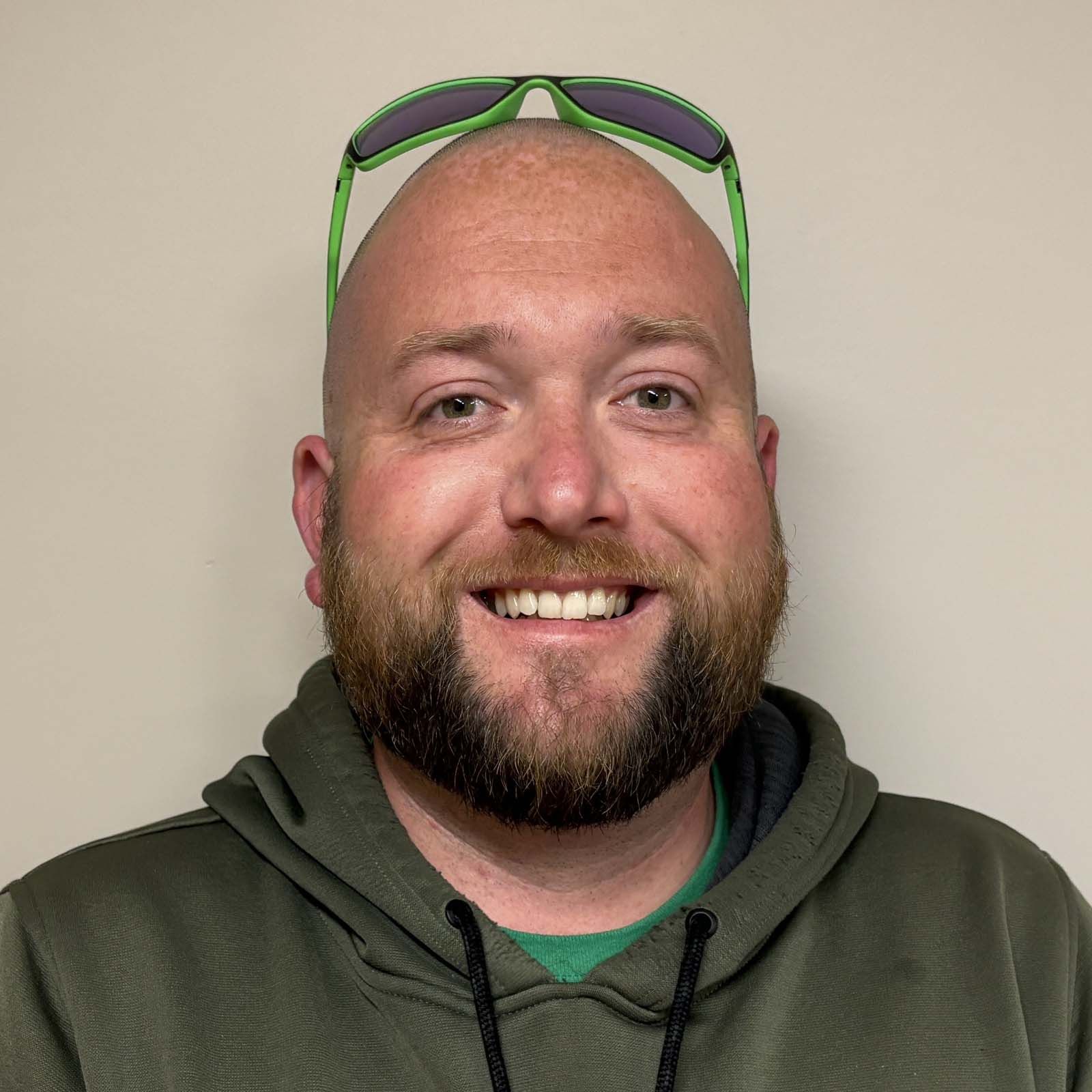 Portrait of a professional staff member wearing a green hoodie and sunglasses resting on the head, standing against a plain light-colored background.