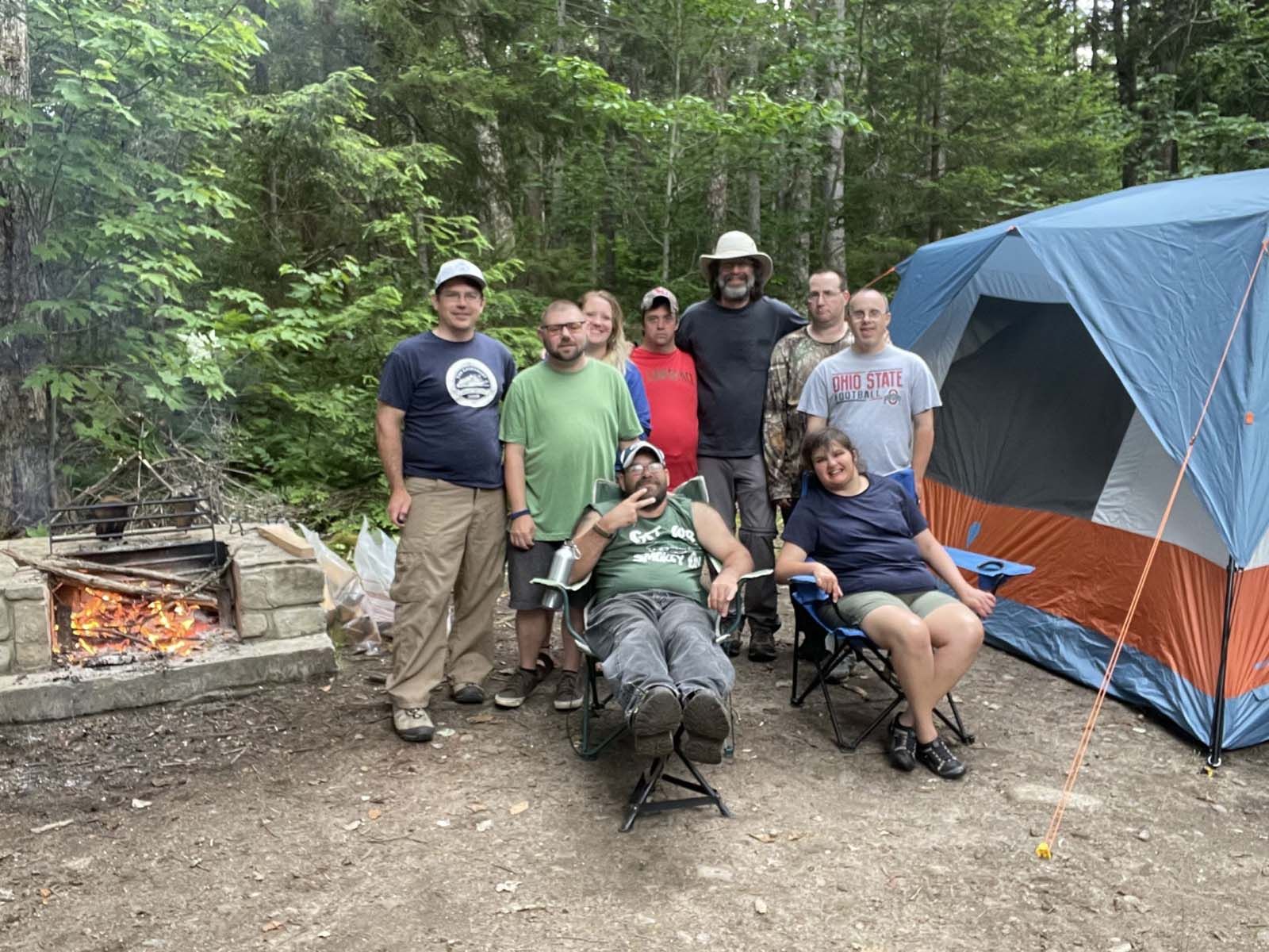 Group of people gathered at a campsite in a wooded area, with several people standing near a tent and picnic area and others seated on camp chairs beside a small fire pit.