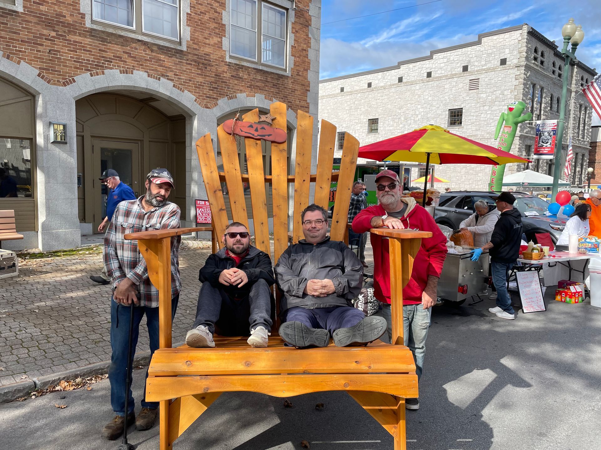Group of people gathered on a street around a large Adirondack style chair, with several people seated in the chair and others standing nearby, buildings and vendor tables visible in the background.