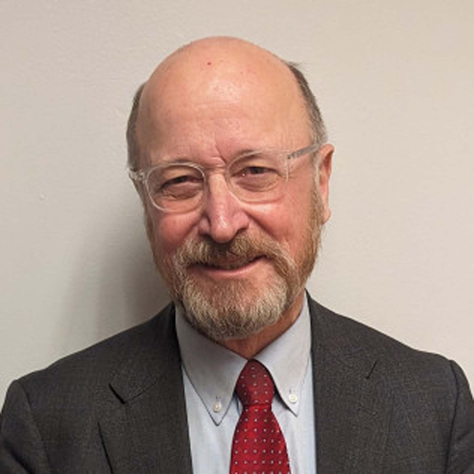Portrait of an executive staff member wearing glasses, a dark suit jacket, a light dress shirt, and a red tie, standing against a plain light-colored background