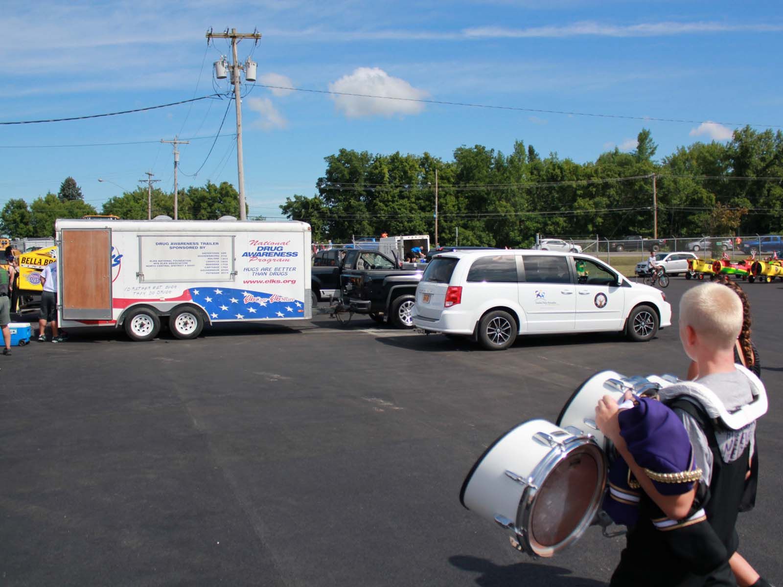 Person wearing a marching band uniform carrying a large drum in a paved outdoor area, with parked vehicles, trailers, and event activity visible in the background.