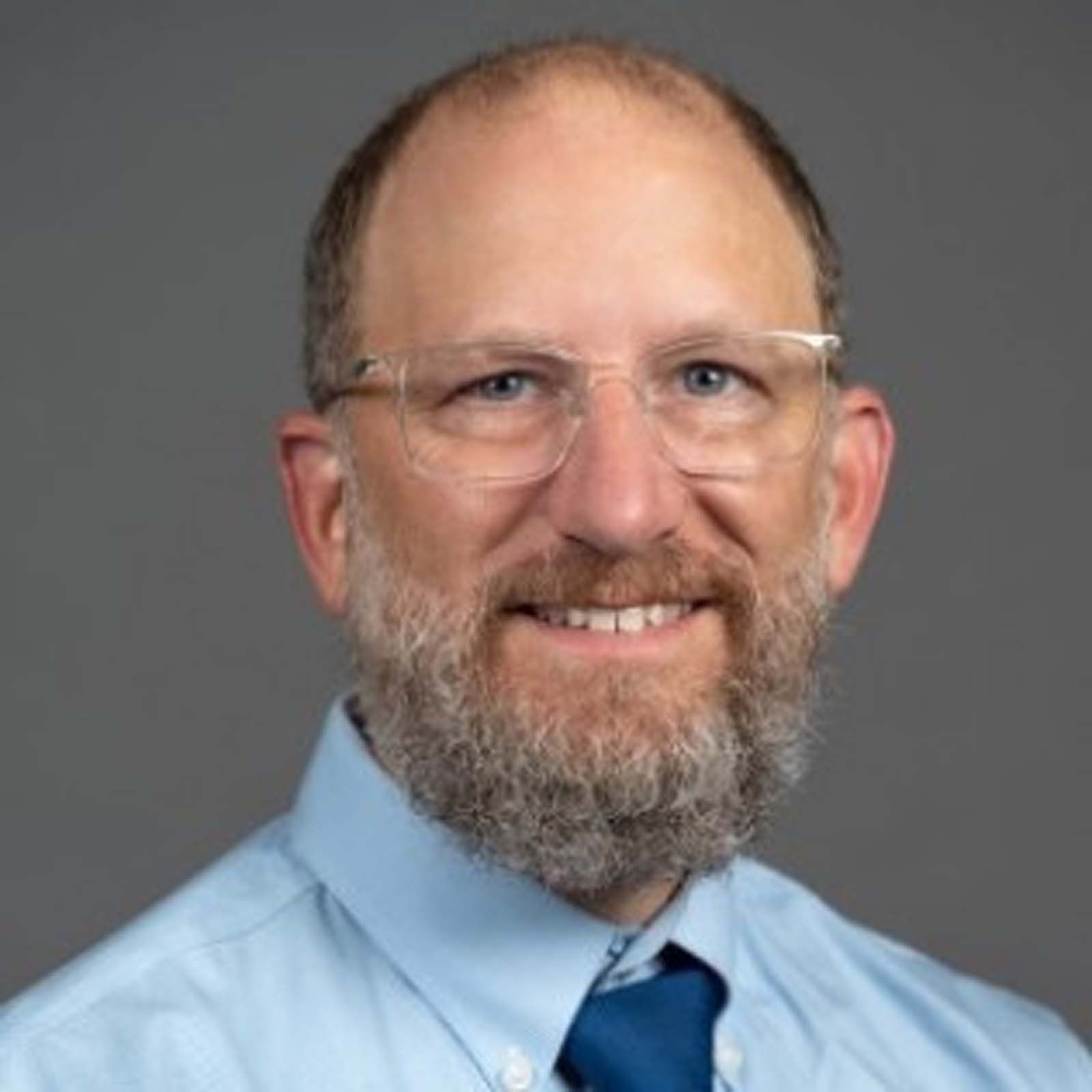 Portrait of a healthcare administrator wearing glasses, a light blue dress shirt, and a blue tie, standing against a plain gray background.