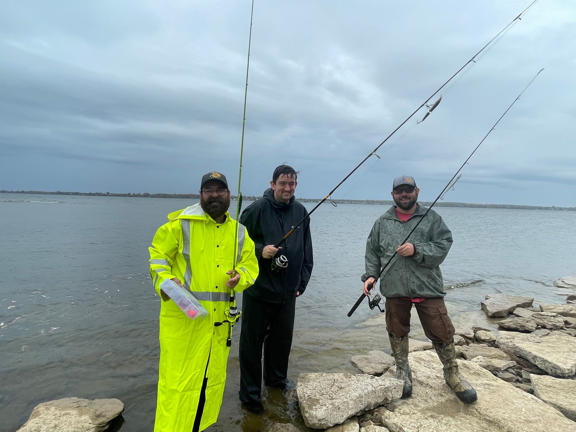 Three people standing on a rocky shoreline by a lake, holding fishing rods, with water and a cloudy sky in the background.