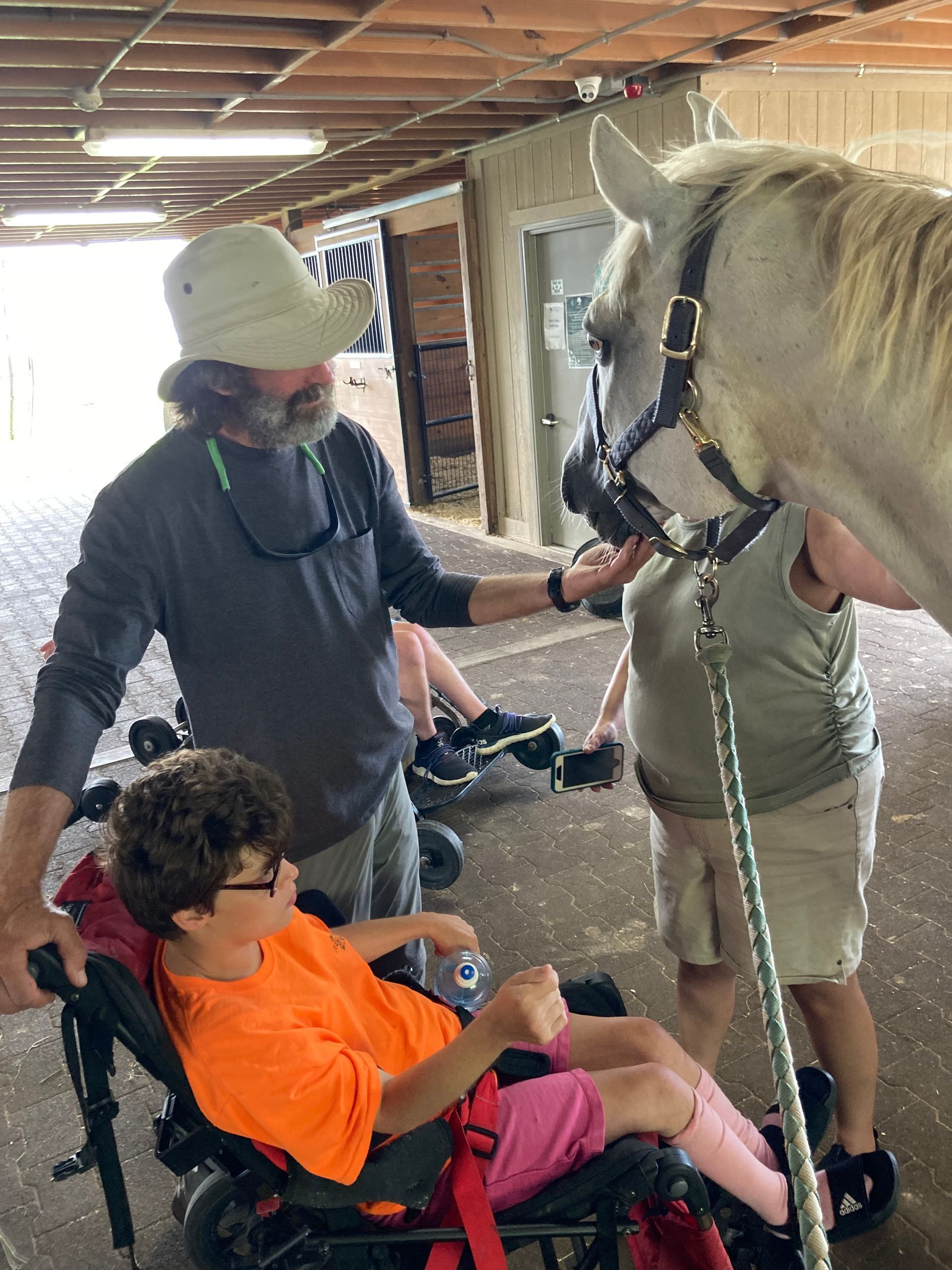 Three people indoors in a stable area interacting with a white horse, with one person seated in a wheelchair and others standing nearby, wooden stalls and equipment visible in the background.