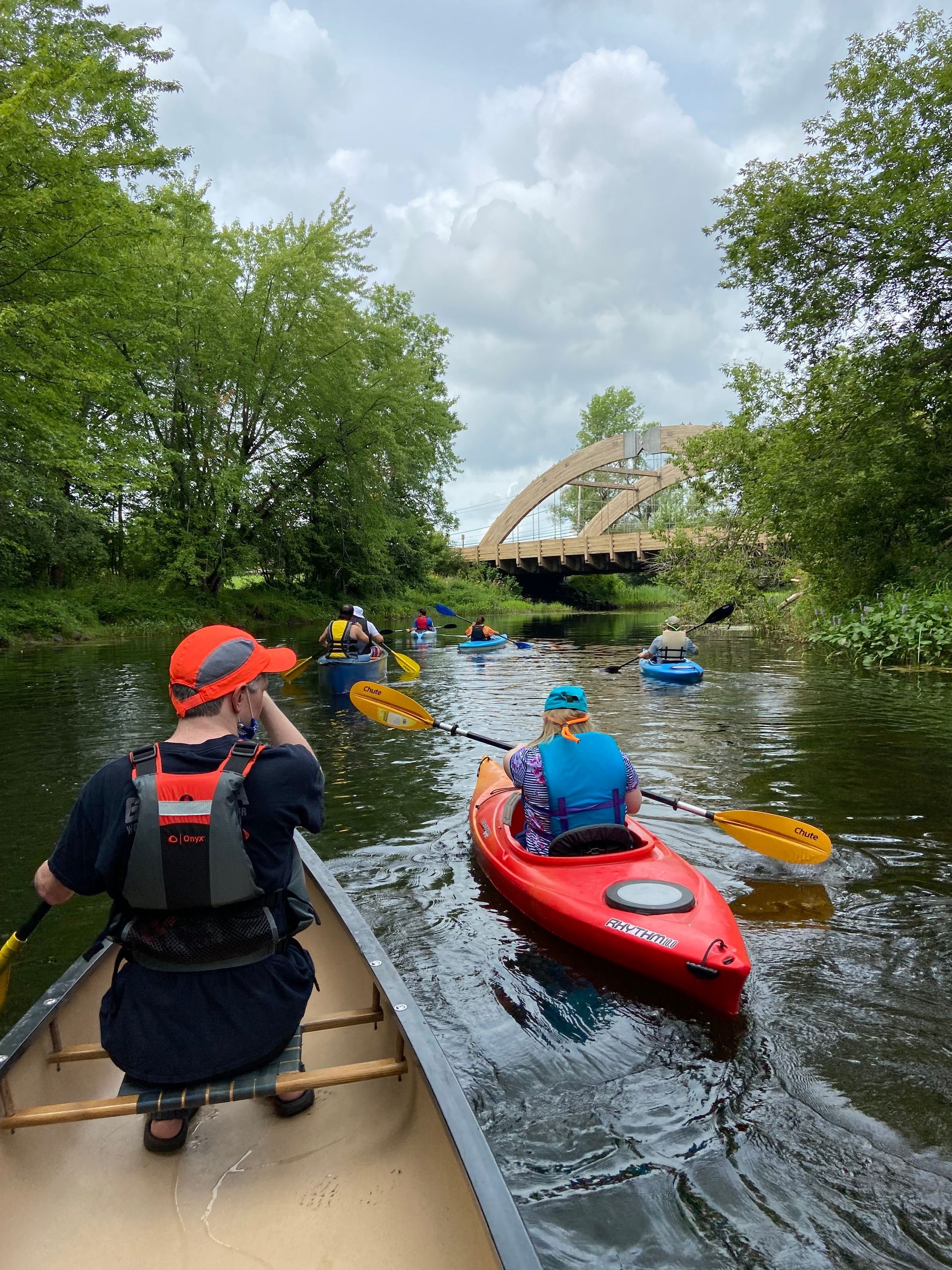 Group of people paddling kayaks and a canoe along a calm river, viewed from behind, with a curved bridge ahead and trees lining both sides of the waterway.