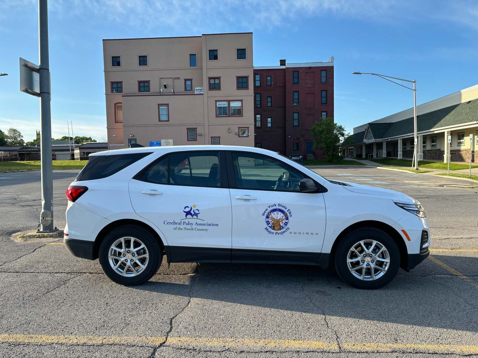 White SUV with organizational logos displayed on the front doors, parked on a paved street with buildings and a blue sky in the background.