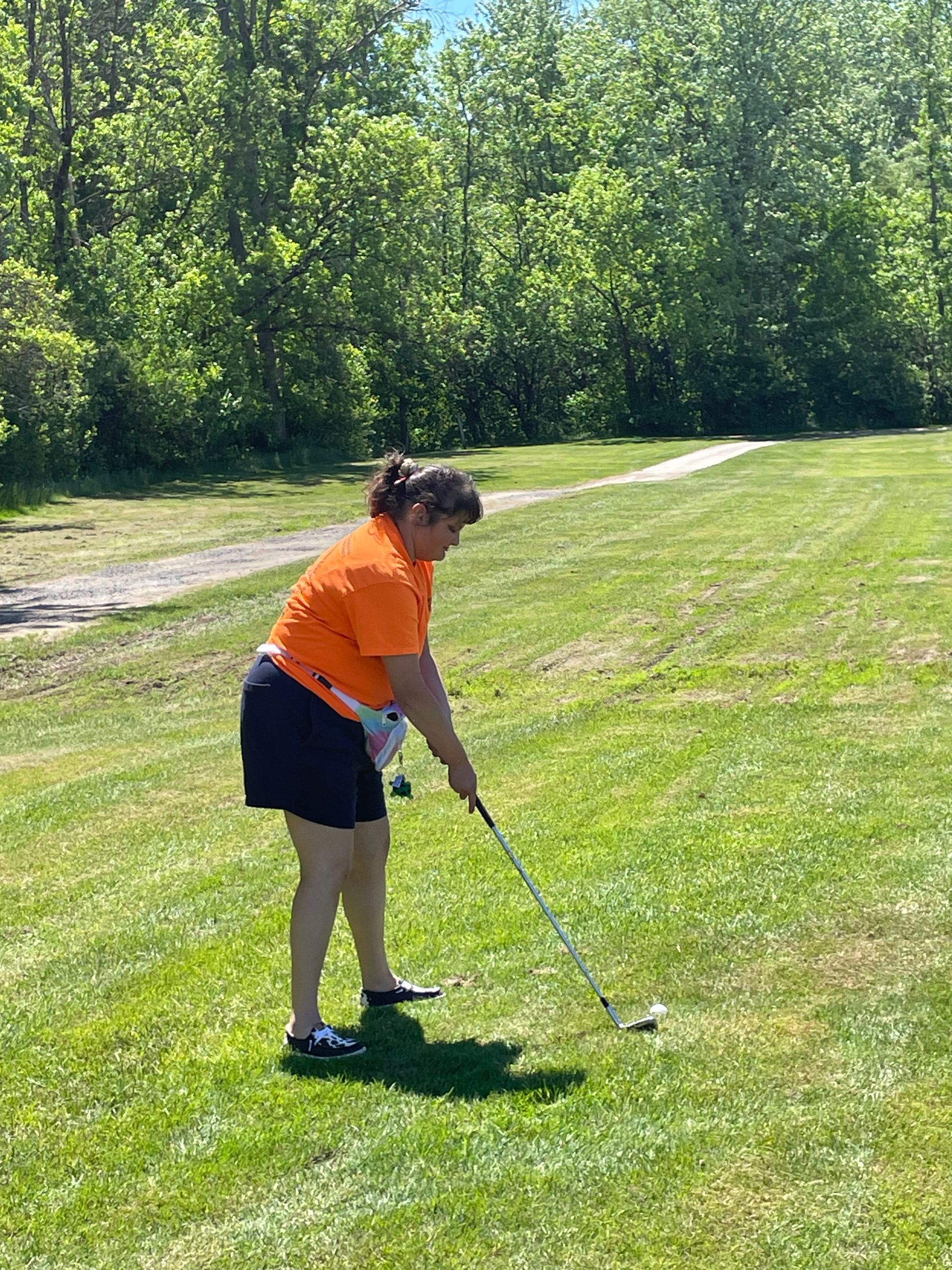 Person standing on a grassy outdoor area holding a golf club and preparing to hit a golf ball, with trees and a walking path visible in the background.