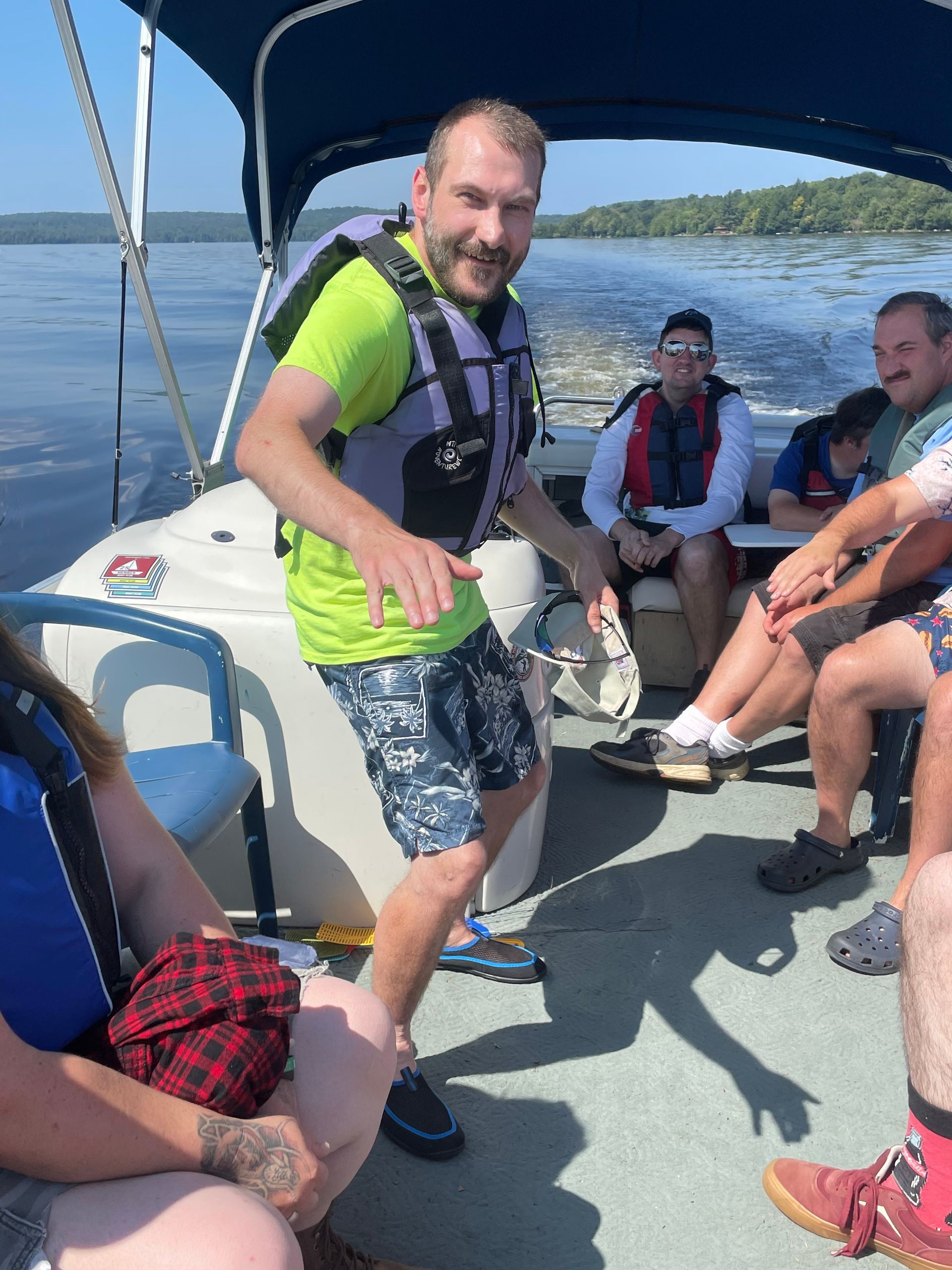 Group of people seated inside a small motorboat on a lake, wearing life vests, with one person standing near the center of the boat and water and shoreline visible in the background.