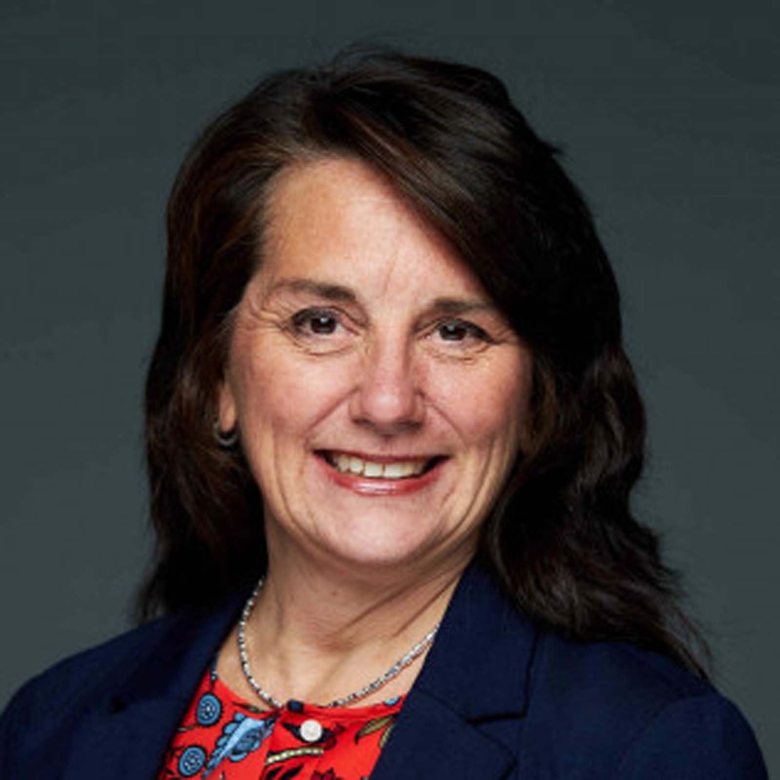Portrait of a senior staff member wearing a dark blazer and a patterned top, standing against a plain gray background.