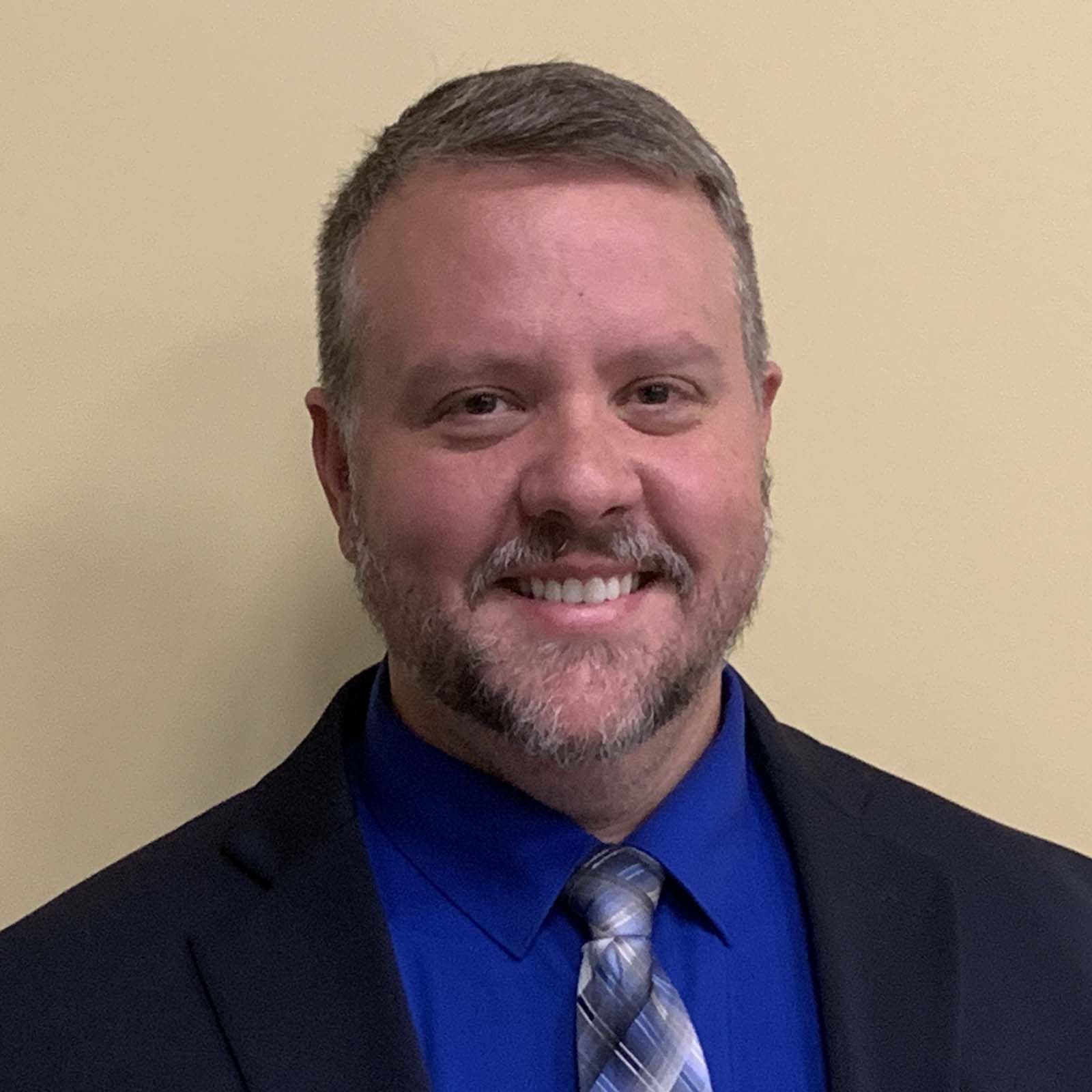 Portrait of a professional staff member wearing a dark suit jacket, a blue dress shirt, and a patterned tie, standing against a plain light colored background