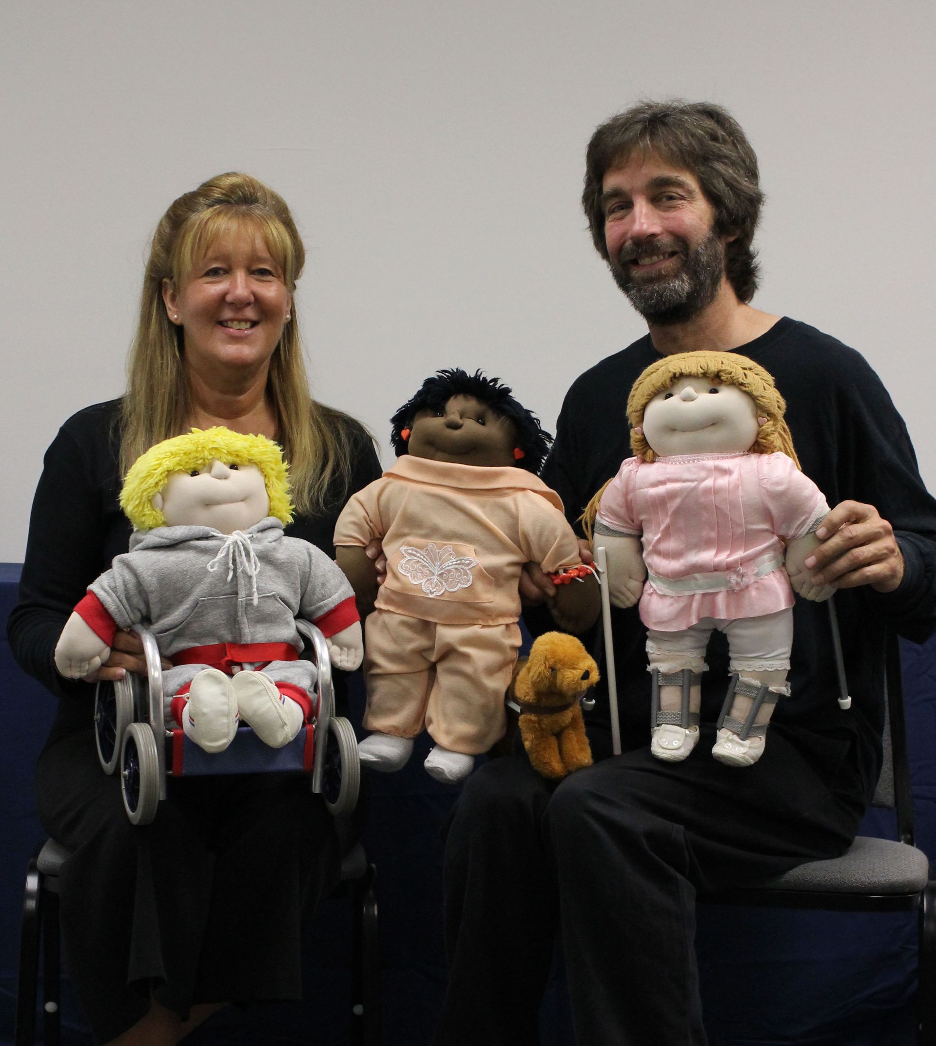 Two people seated indoors holding several cloth dolls of different sizes and styles, with one in a wheelchair and a plain wall visible in the background.