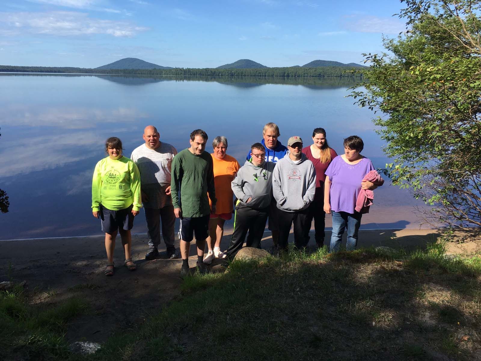 Group of people standing together on a grassy shoreline beside a calm lake, with trees in the foreground and mountains visible across the water in the background.