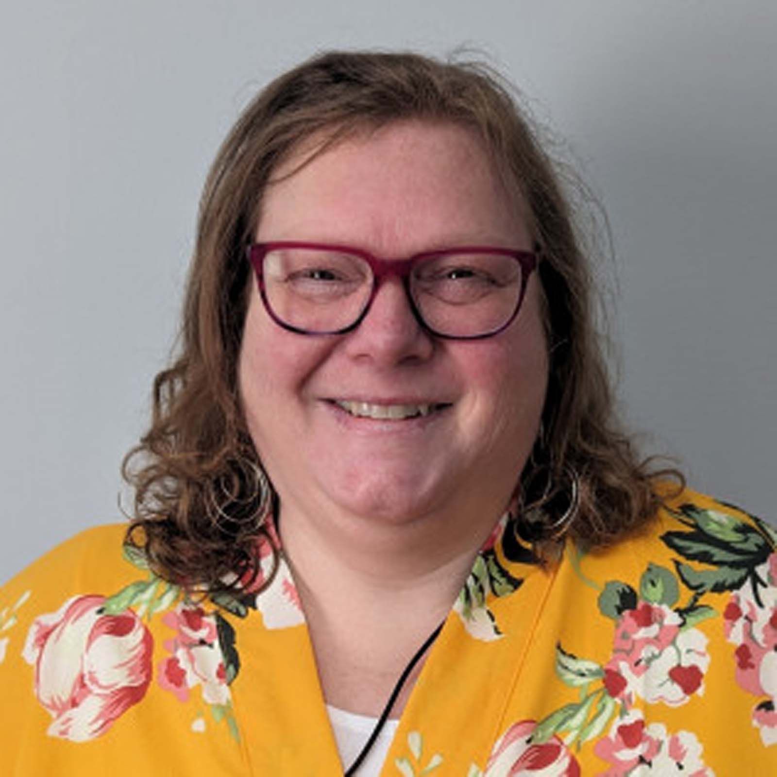 Portrait of a professional staff member wearing glasses and a yellow floral patterned top, standing against a plain light colored background.
