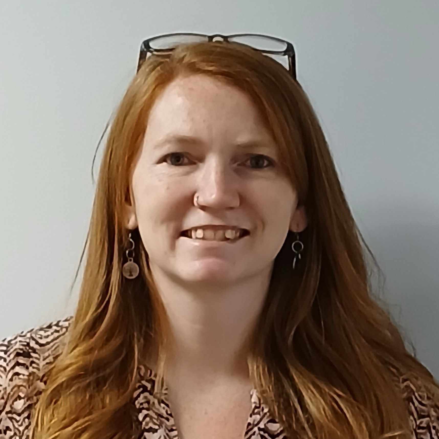 Portrait of a professional staff member wearing a patterned top and glasses resting on the head, standing against a plain light colored background.