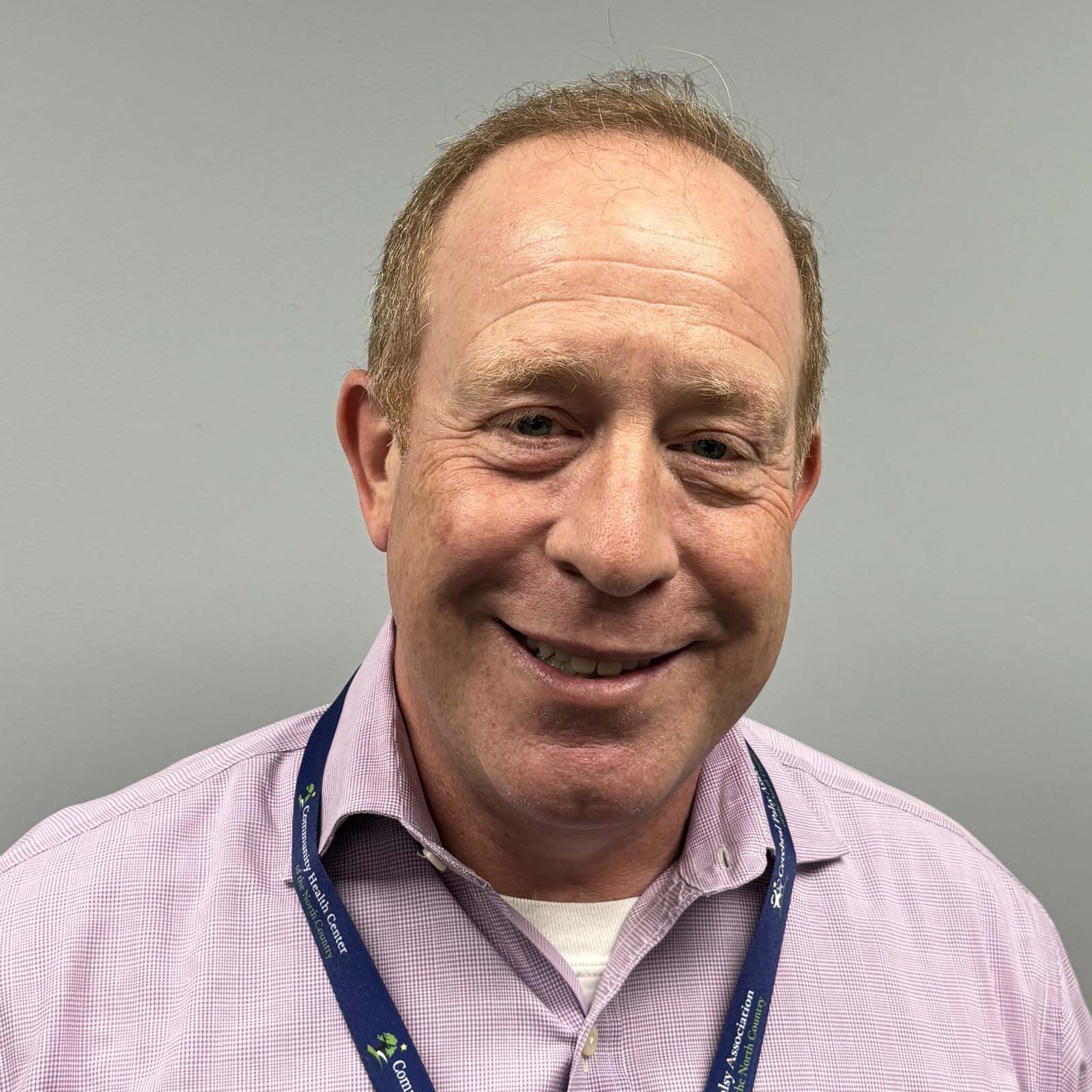 Portrait of a professional staff member wearing a light purple button down shirt and a lanyard, standing against a plain gray background.