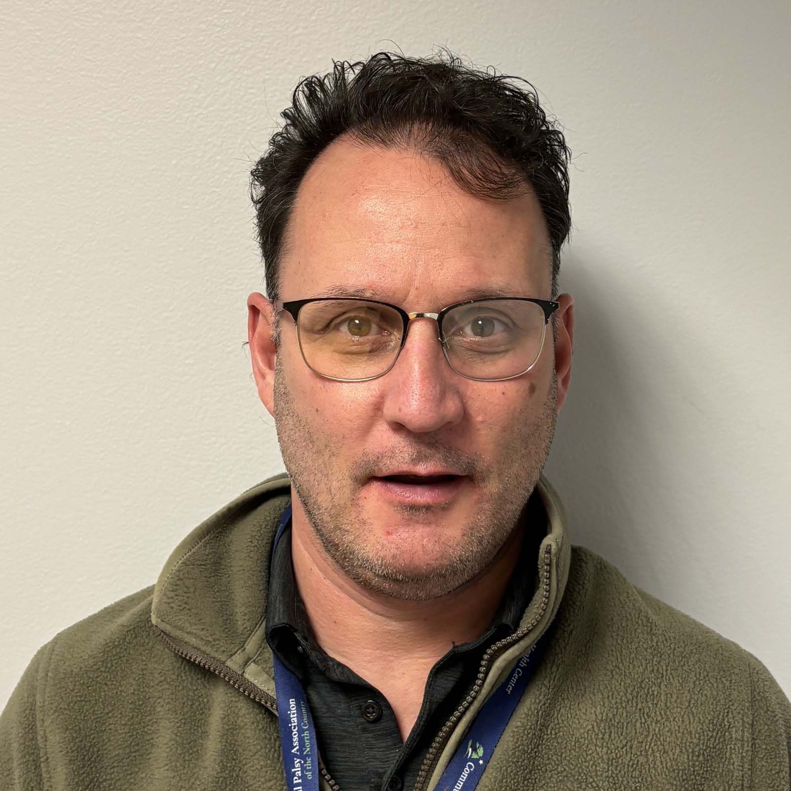 Portrait of a professional staff member wearing glasses, a green fleece jacket, and a lanyard, standing against a plain light colored wall.