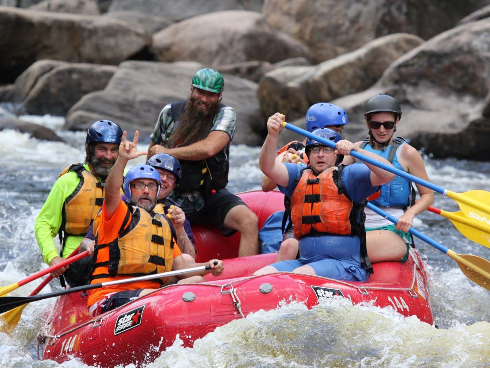 Group of people wearing helmets and life vests paddling a red inflatable raft through fast-moving river water, with large rocks visible in the background.