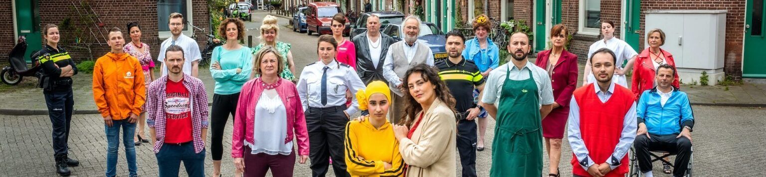 Een groep mensen staat op een straat met kinderkopjes. De meesten kijken naar de camera. Op de achtergrond zijn gebouwen en begroeiing te zien.