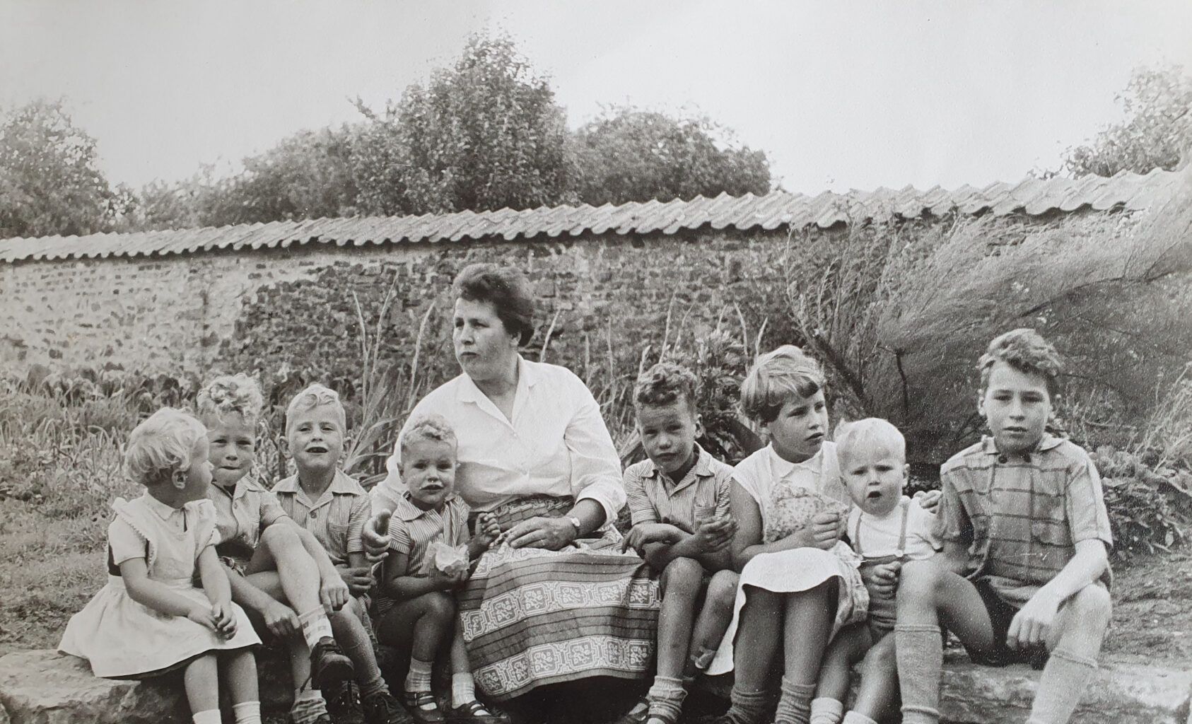 Een vrouw zit met een groep kinderen voor een stenen muur en begroeiing.