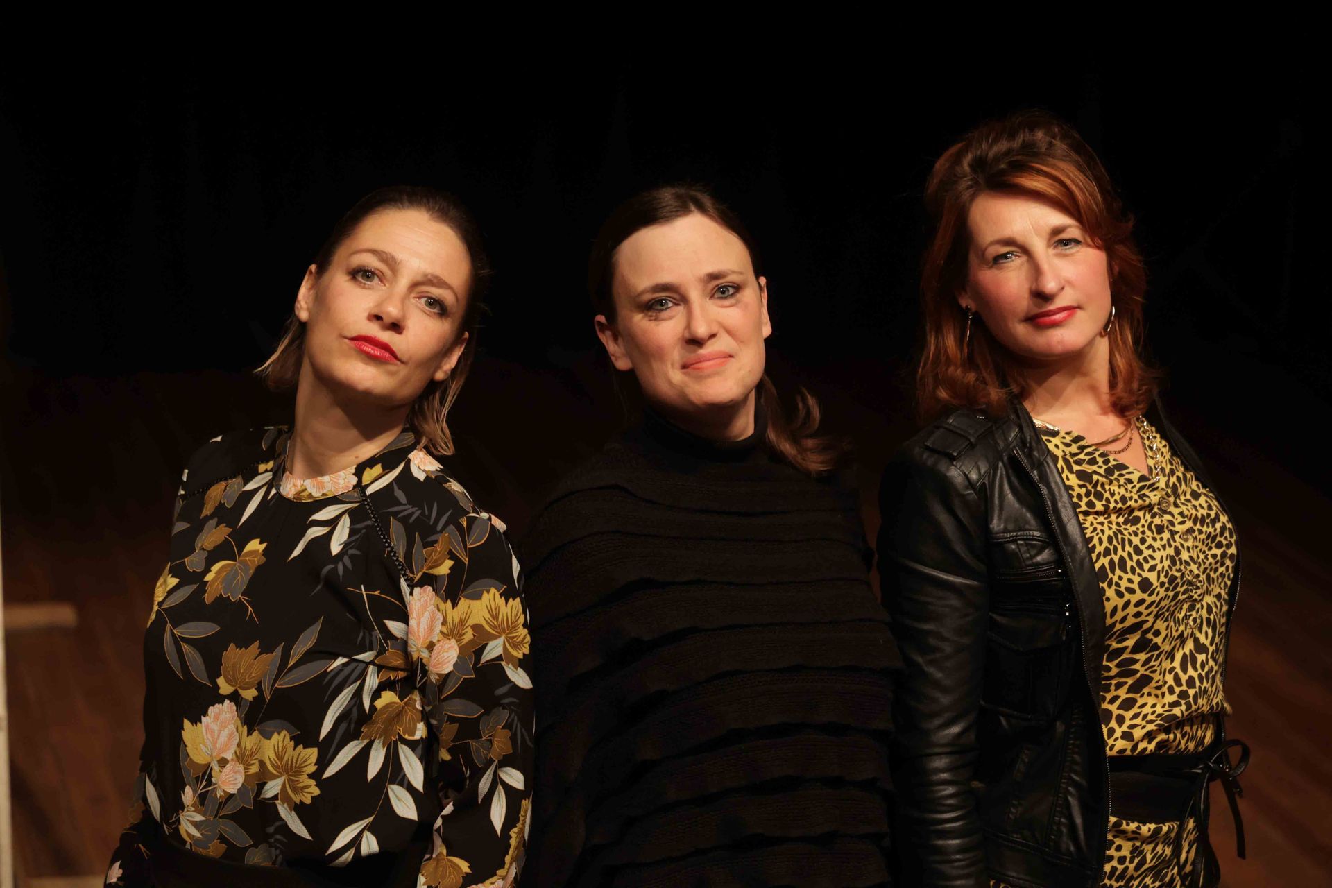 Drie vrouwen poseren voor een foto op een podium. De ene draagt ​​een jurk met bloemenprint, de andere een zwarte top en de derde een leren jasje over een jurk met luipaardprint.