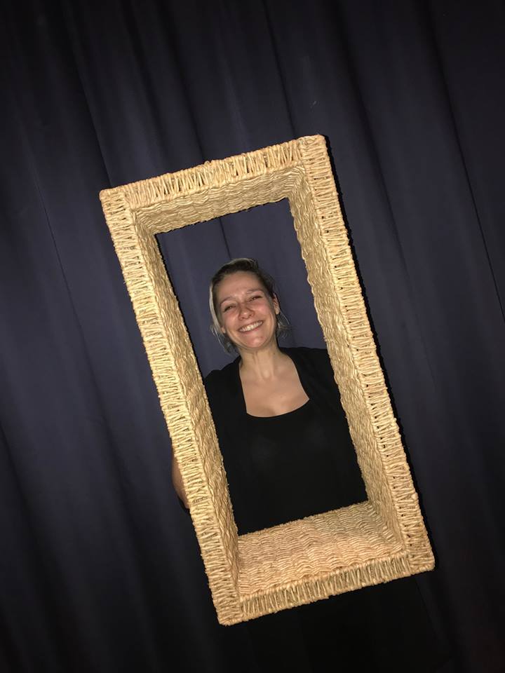 Een vrouw glimlacht terwijl ze een rechthoekig frame met textuur vasthoudt; op de achtergrond een blauw gordijn.