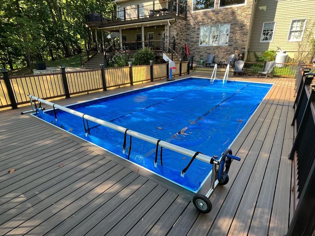 A rectangular swimming pool covered in blue with a rolling cover on a wooden deck. A house is in the background.